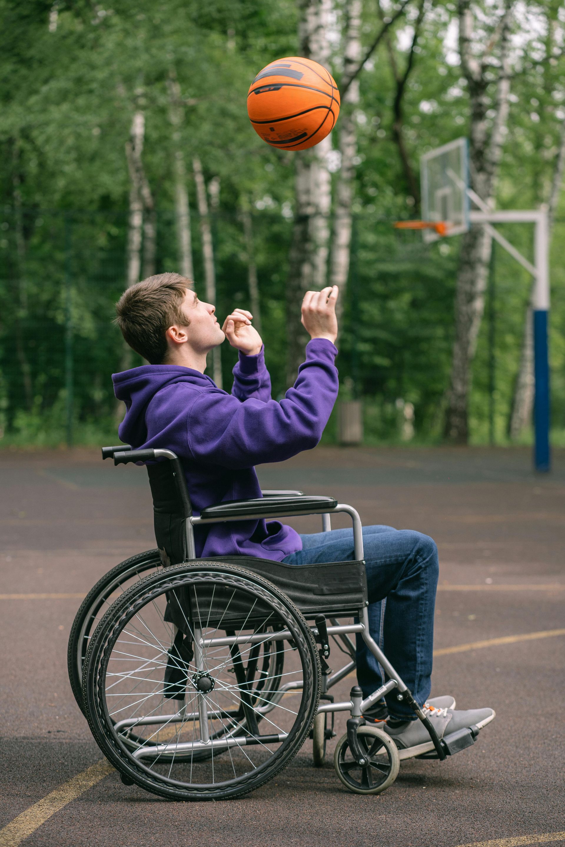 Man in wheelchair tossing a basketball toward a hoop on a court. He wears a purple hoodie and jeans.