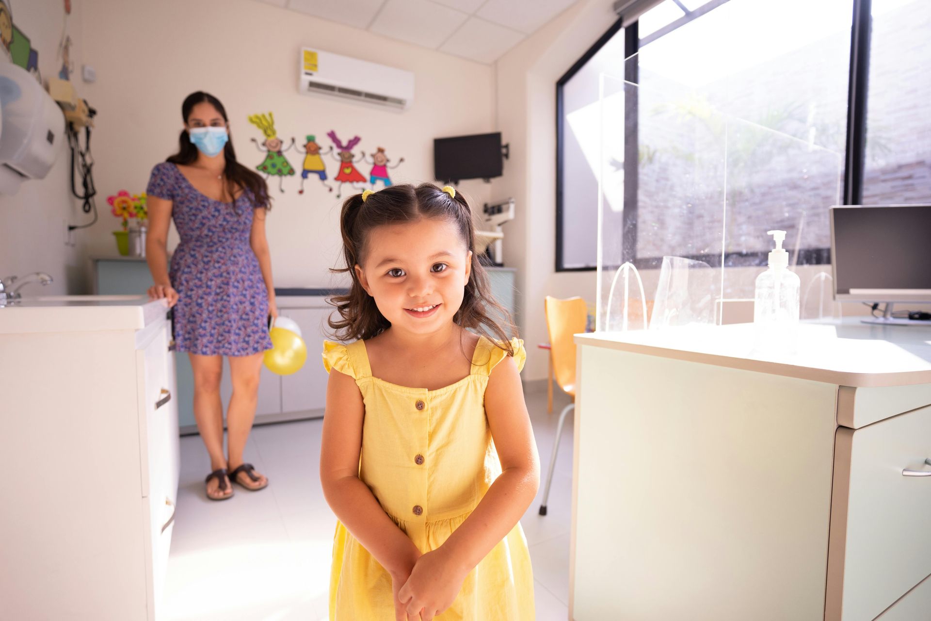 Smiling child in yellow dress; woman with mask in background. Bright room.