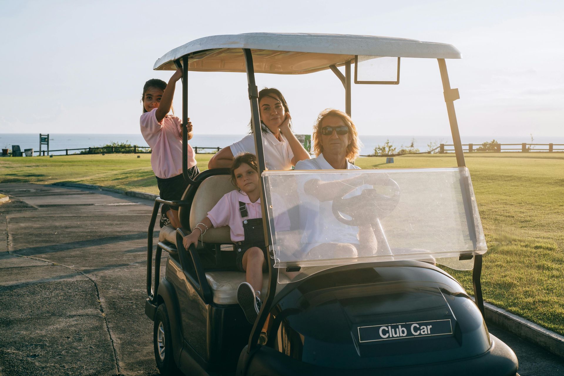 Four people in a golf cart on a grassy field near the ocean.