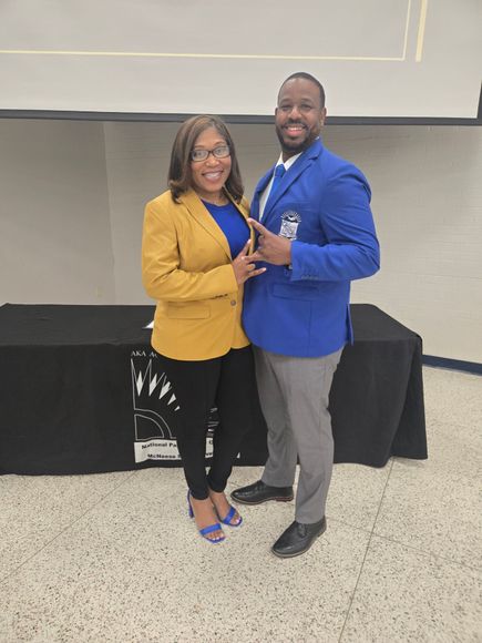 Woman in yellow blazer and man in blue blazer stand together. Black table with banner.