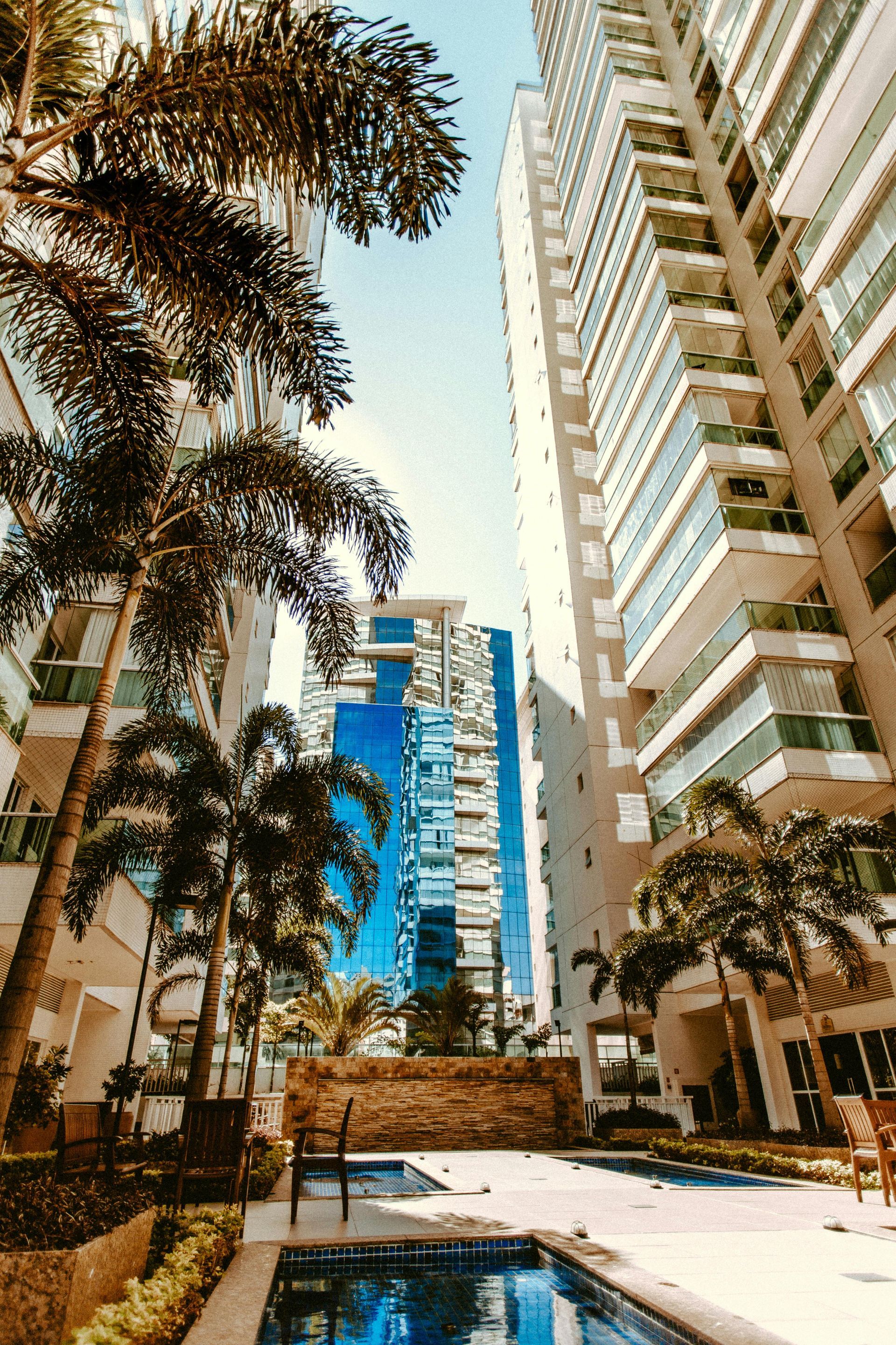 Pool area between tall white apartment buildings, palm trees, and a blue glass tower.