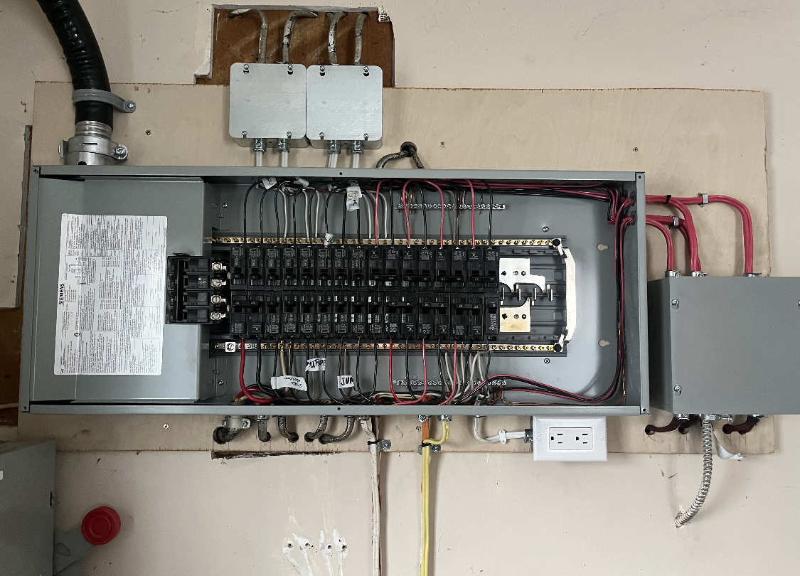 Electrical panel with numerous breakers, wires, and conduits mounted on a wall. A receptacle is visible below.