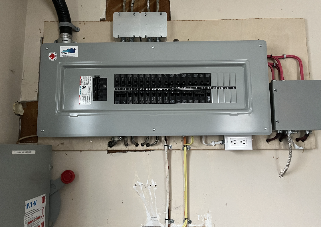Electrical panel with circuit breakers mounted on a wall. Wires and conduit are visible.