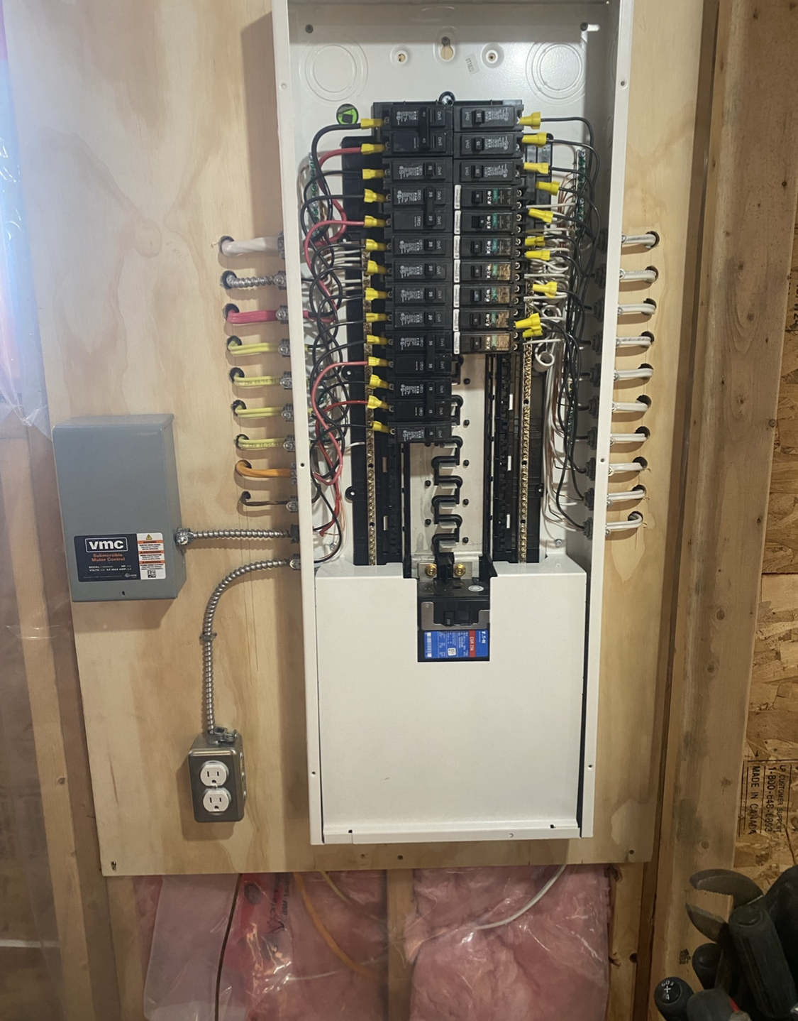 Electrical panel with numerous circuit breakers and wiring mounted on a wooden wall.