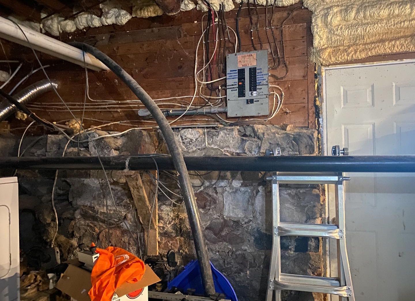 Basement with exposed stone wall, electrical panel, pipes, and ladder.