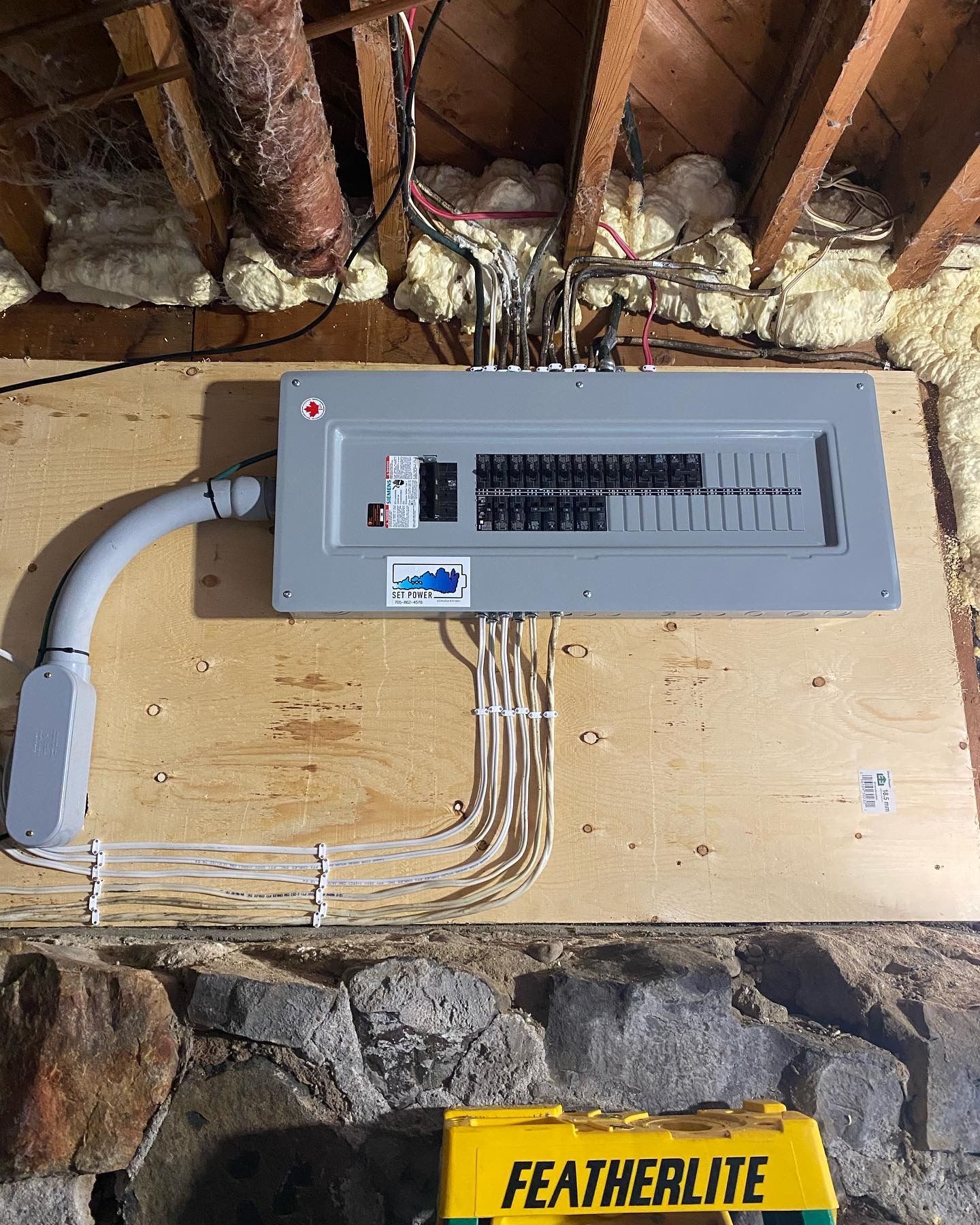 Electrical panel mounted on wood, with conduit, cables, and wires against a stone wall.