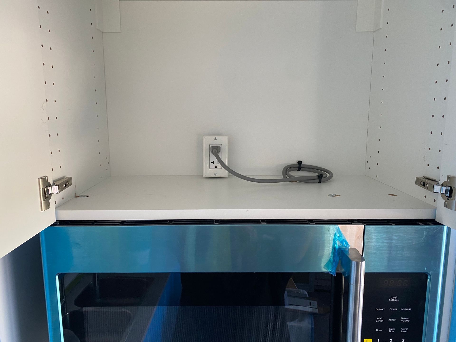 White cabinet interior with a microwave below and an electrical outlet and cord on the shelf.