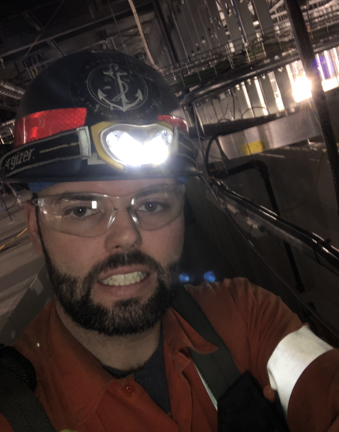 Man in hard hat and safety glasses with a headlamp, smiling in an industrial setting.