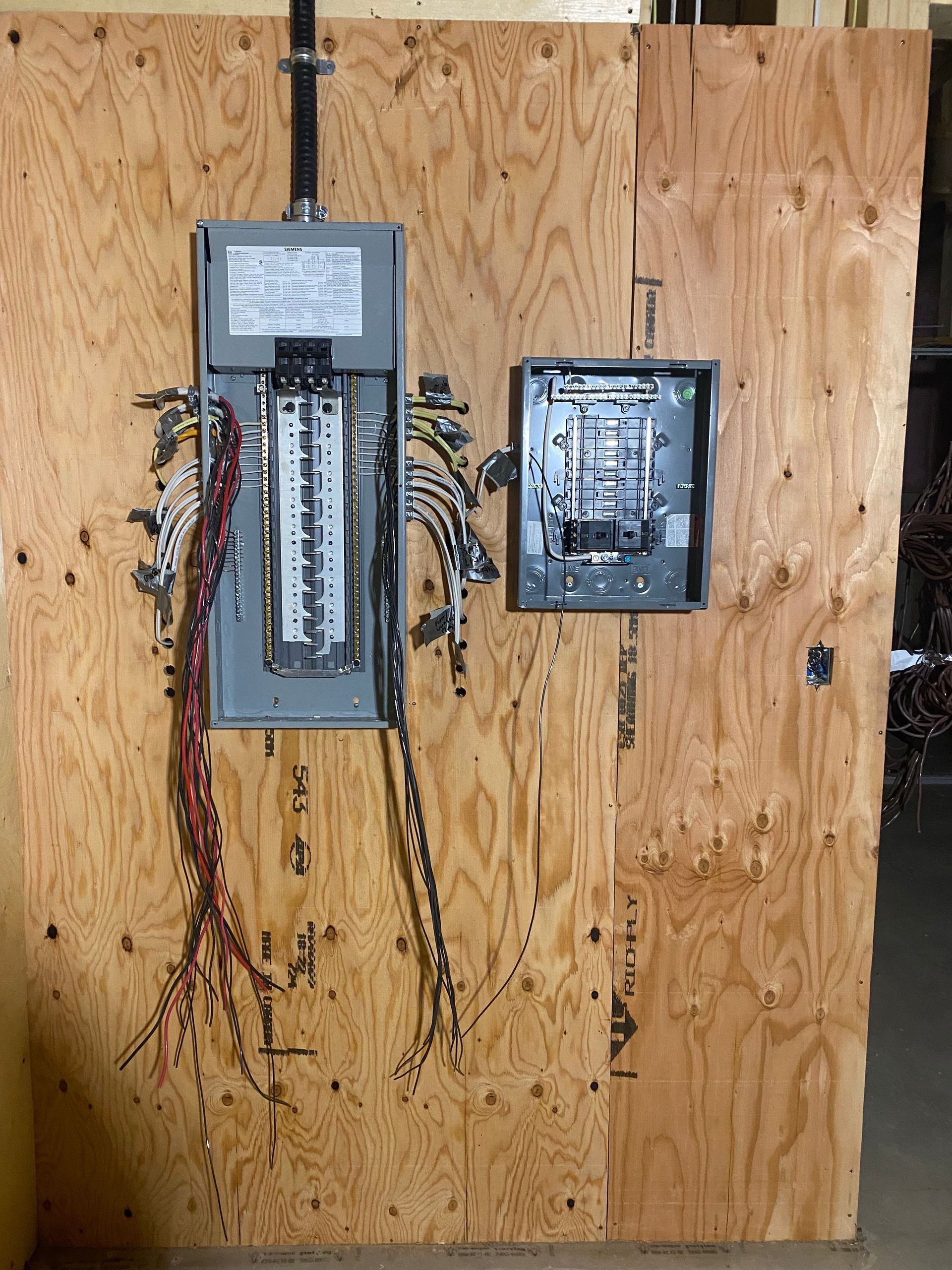 Electrical panel mounted on a plywood board with exposed wiring. Includes a breaker box and a smaller subpanel.