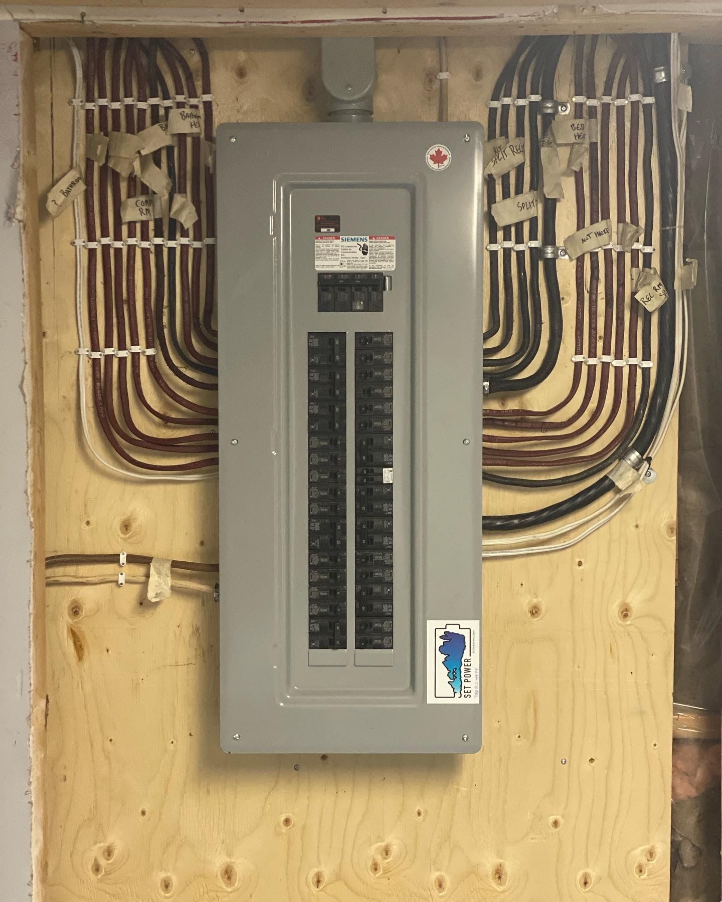 Electrical panel mounted on a wood wall with numerous wires labeled and secured.