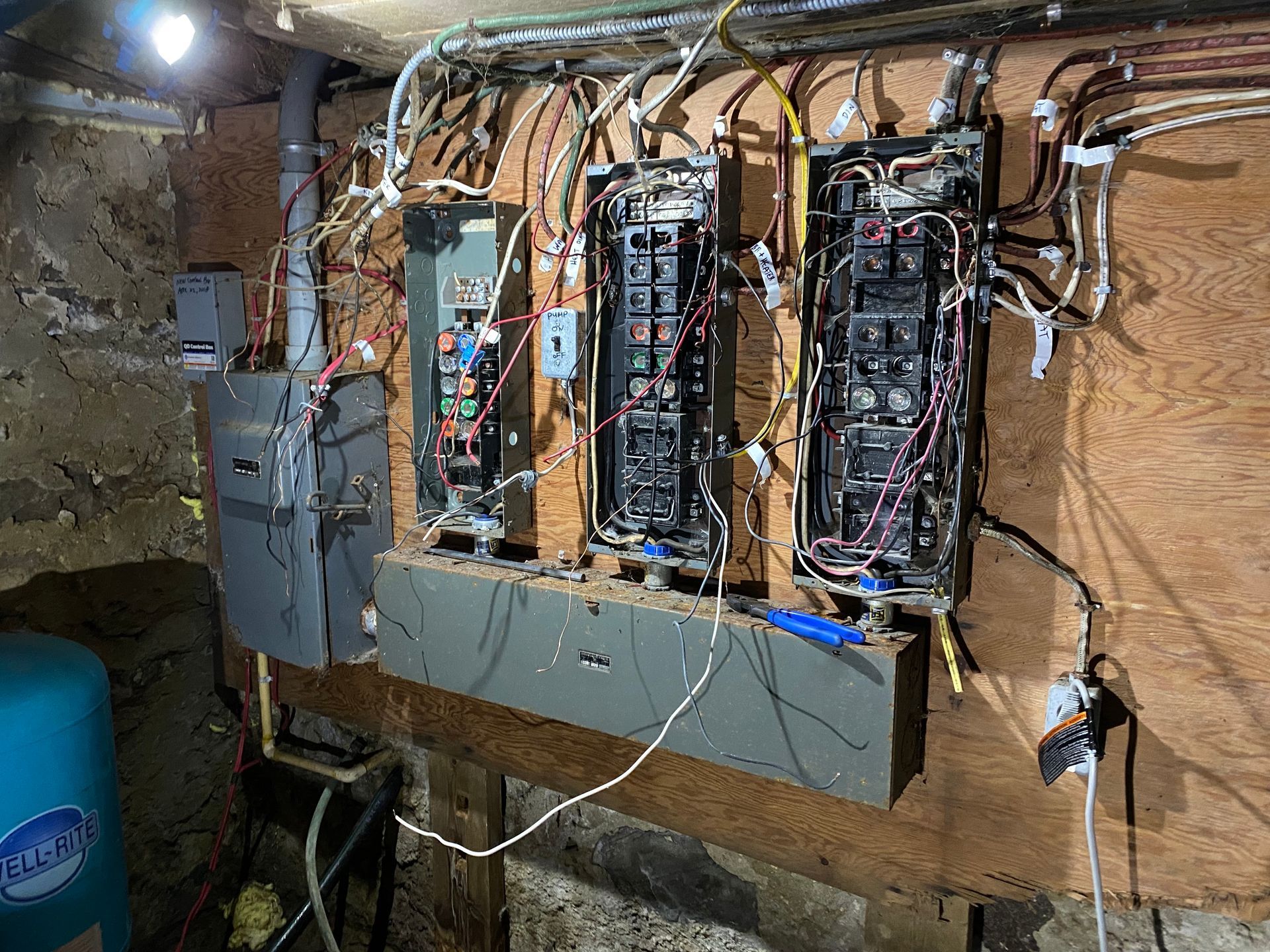 Electrical panels mounted on a wooden wall in a basement, with wires and conduits visible.