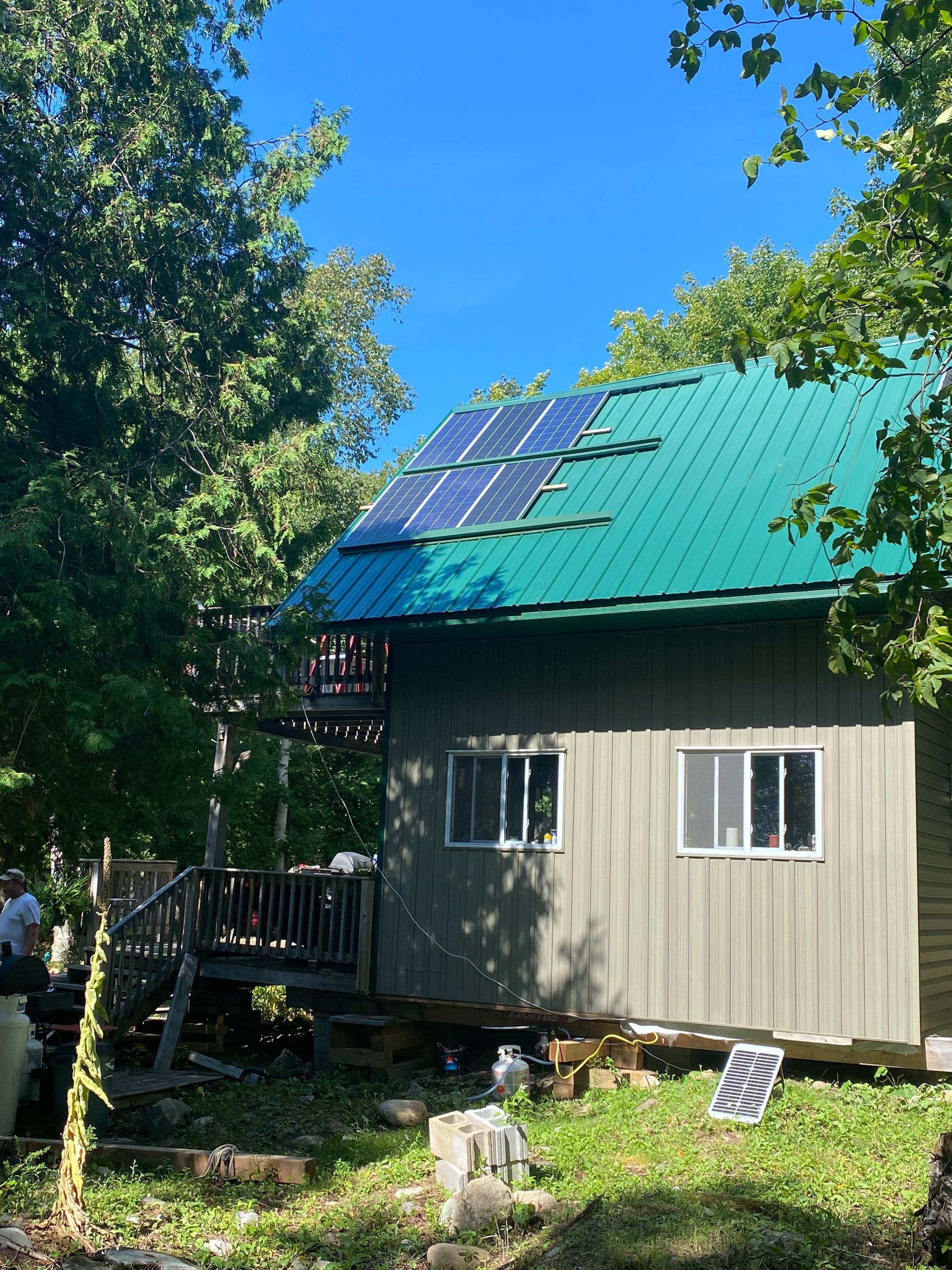 Cabin with green metal roof, solar panels, surrounded by trees under a clear blue sky.