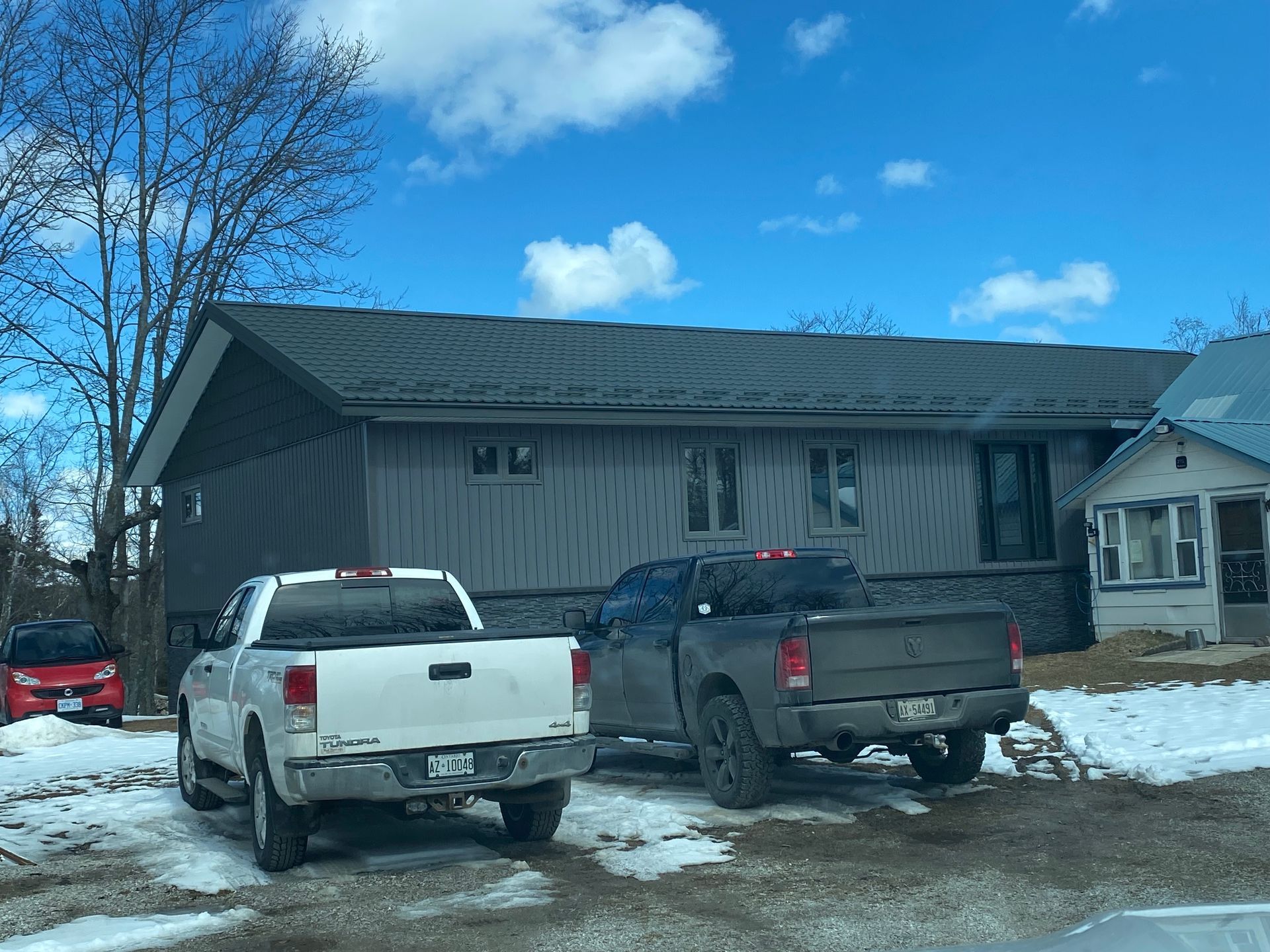 Two pickup trucks parked in front of a gray house on a snowy day with blue sky.