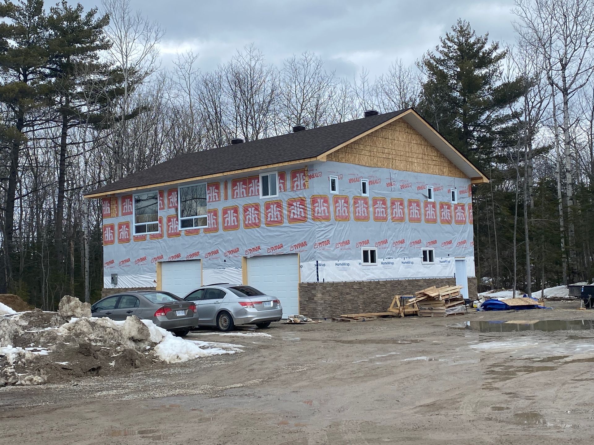 Two-story house under construction with a dark roof and a stone facade, surrounded by trees and a dirt driveway.