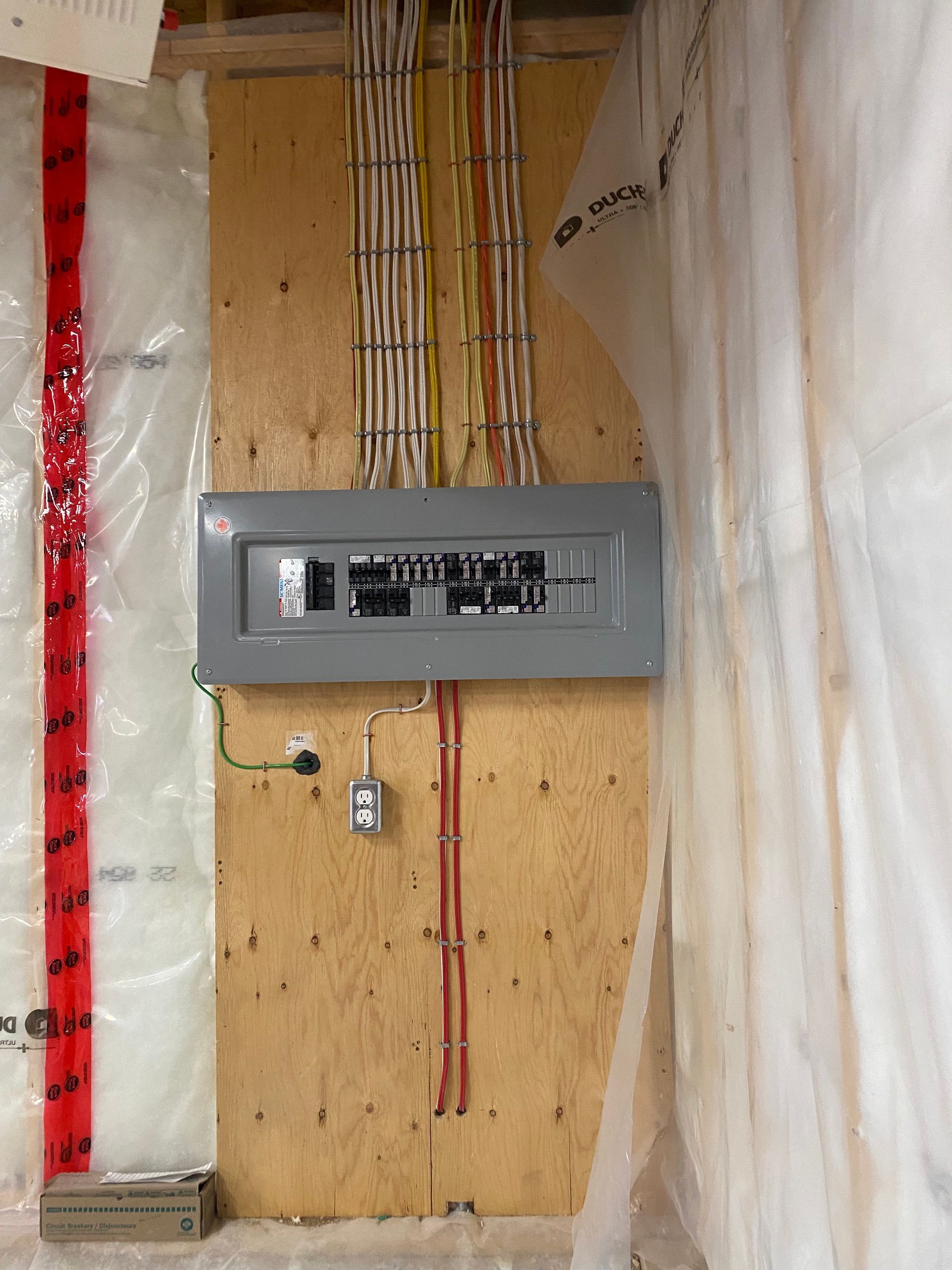 Electrical panel on plywood wall, with wiring. Red and yellow wires bundled above, conduit below. Insulation and plastic sheeting present.