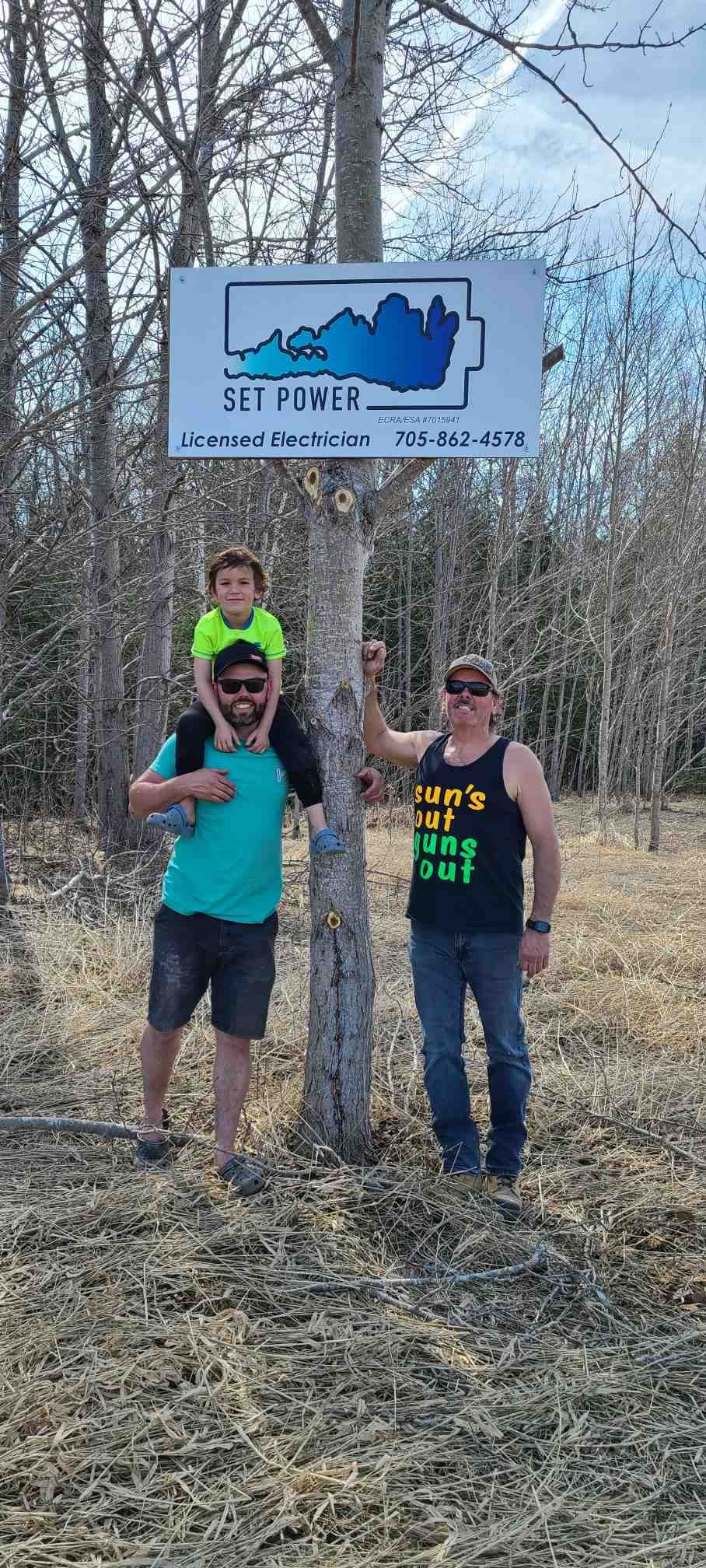 Three people stand by a sign. A man holds a child on his shoulders. Another man leans on a tree. The sign has a blue outline of Kentucky.