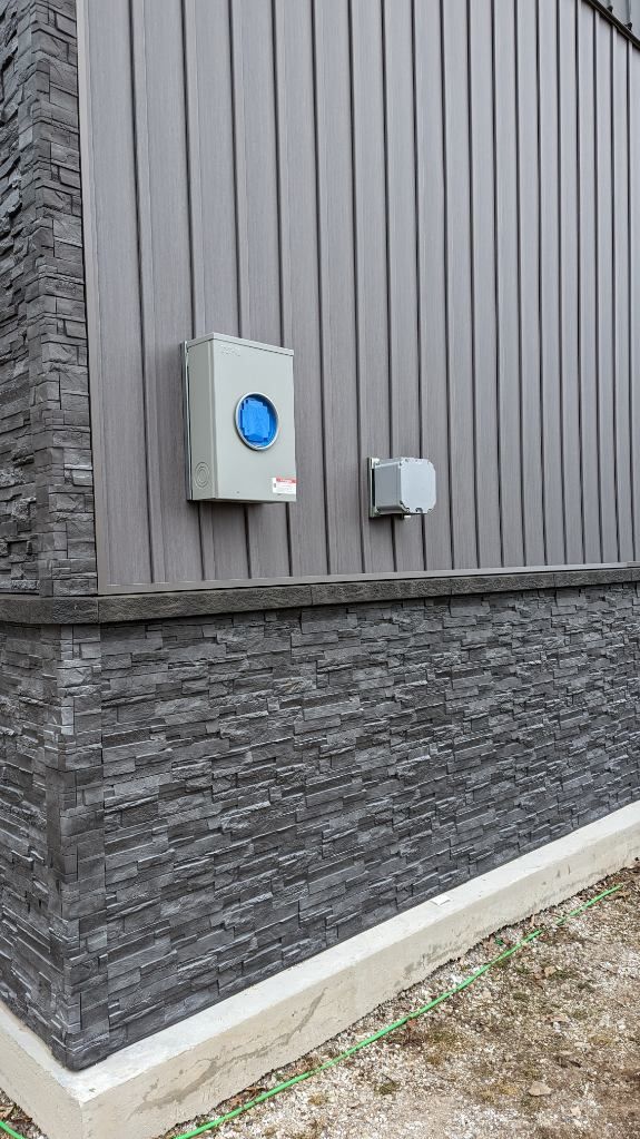 Gray electrical boxes on a building's exterior. Vertical siding and textured stone foundation.