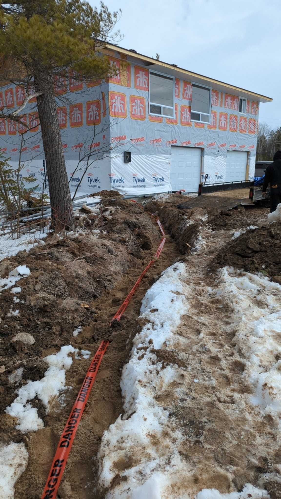 Construction site: house with wood siding, trench with orange electrical conduit, snow and dirt.