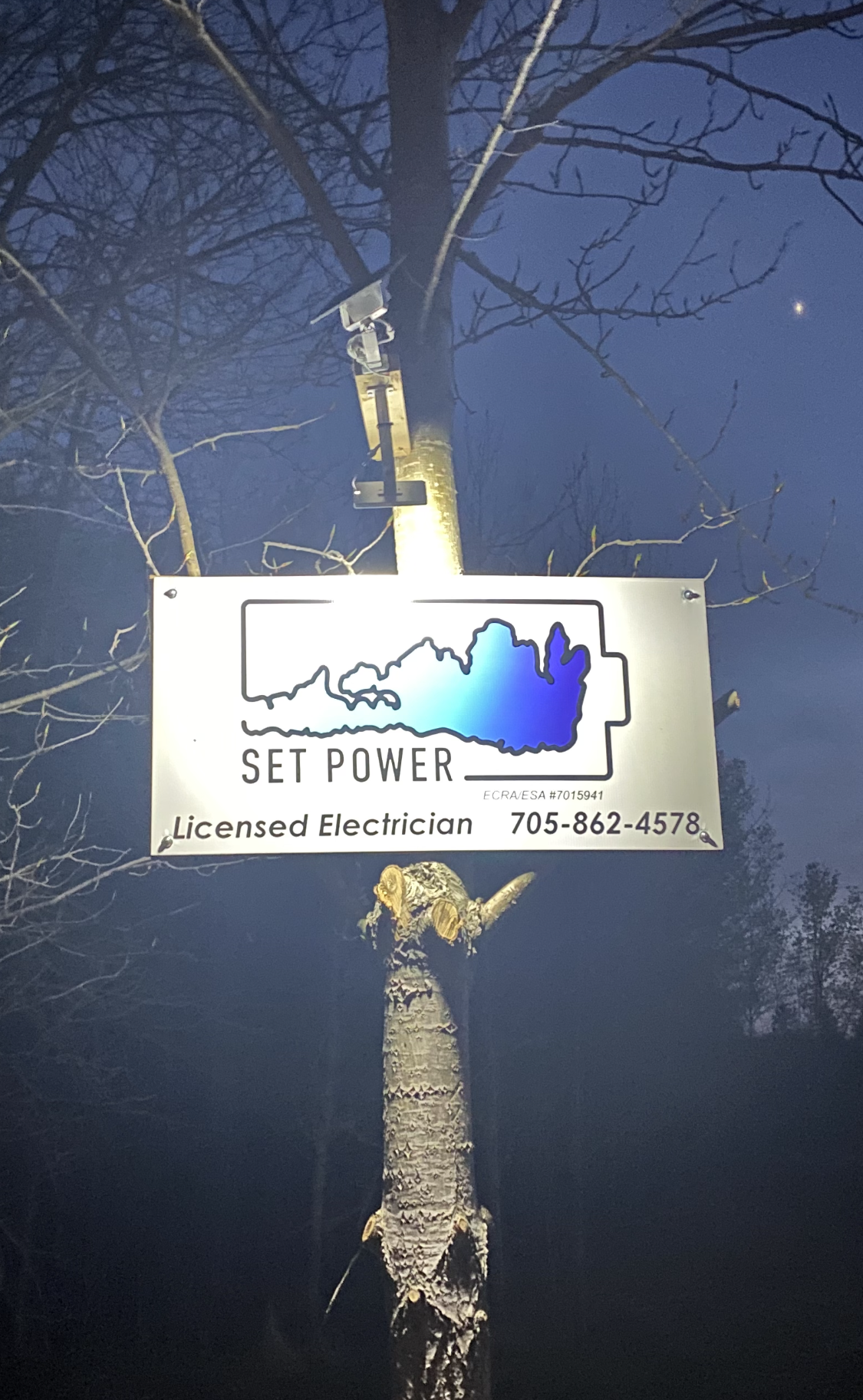 Sign for Set Power, licensed electrician, mounted on a tree; sign has battery graphic with city skyline.