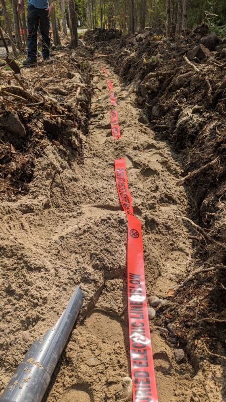 Trench with red markers, ready for electrical work. Man’s legs visible. Outdoors, dirt, trees.