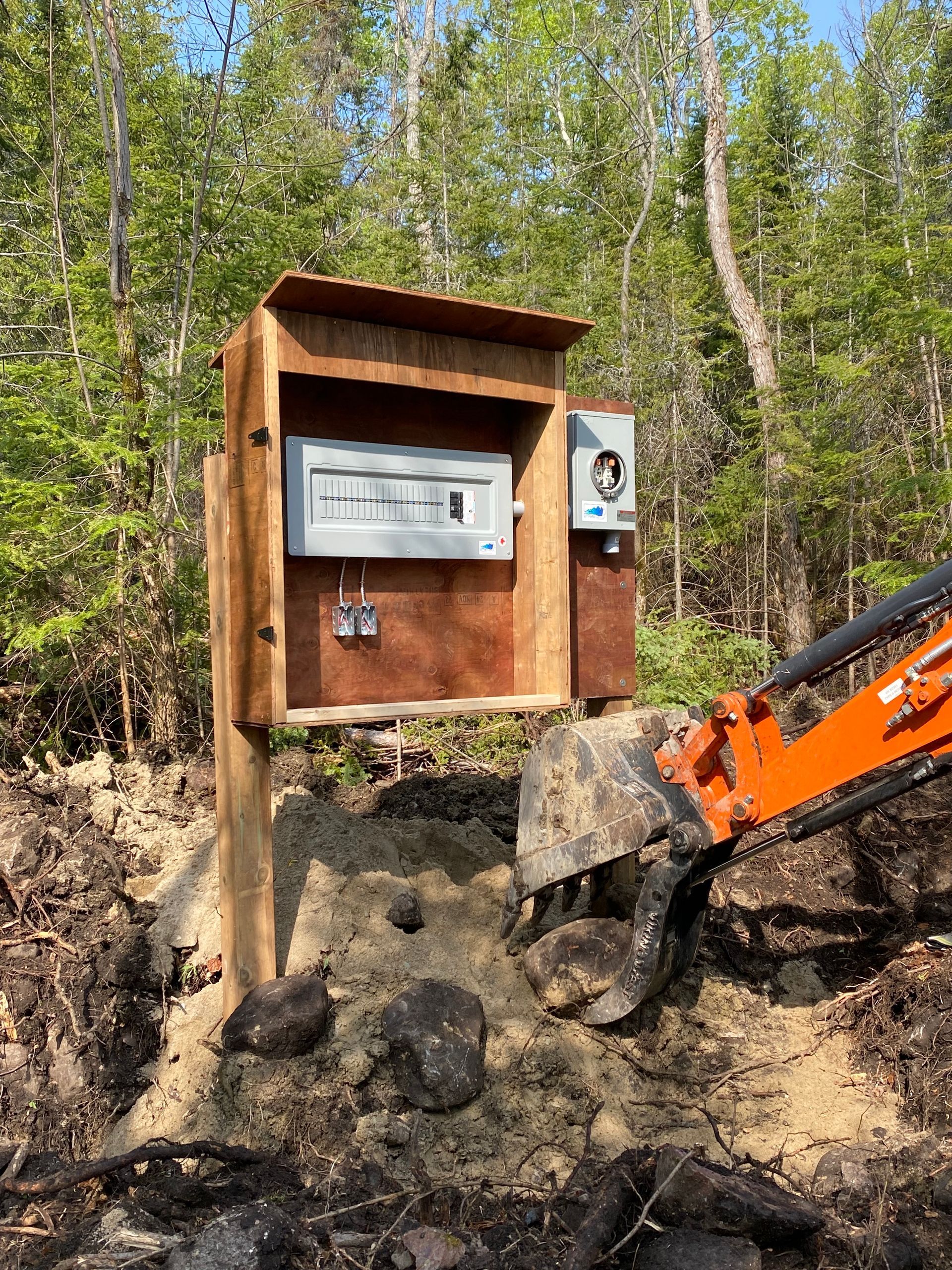 An electrical panel on a wooden post in a wooded area being worked on by an excavator.