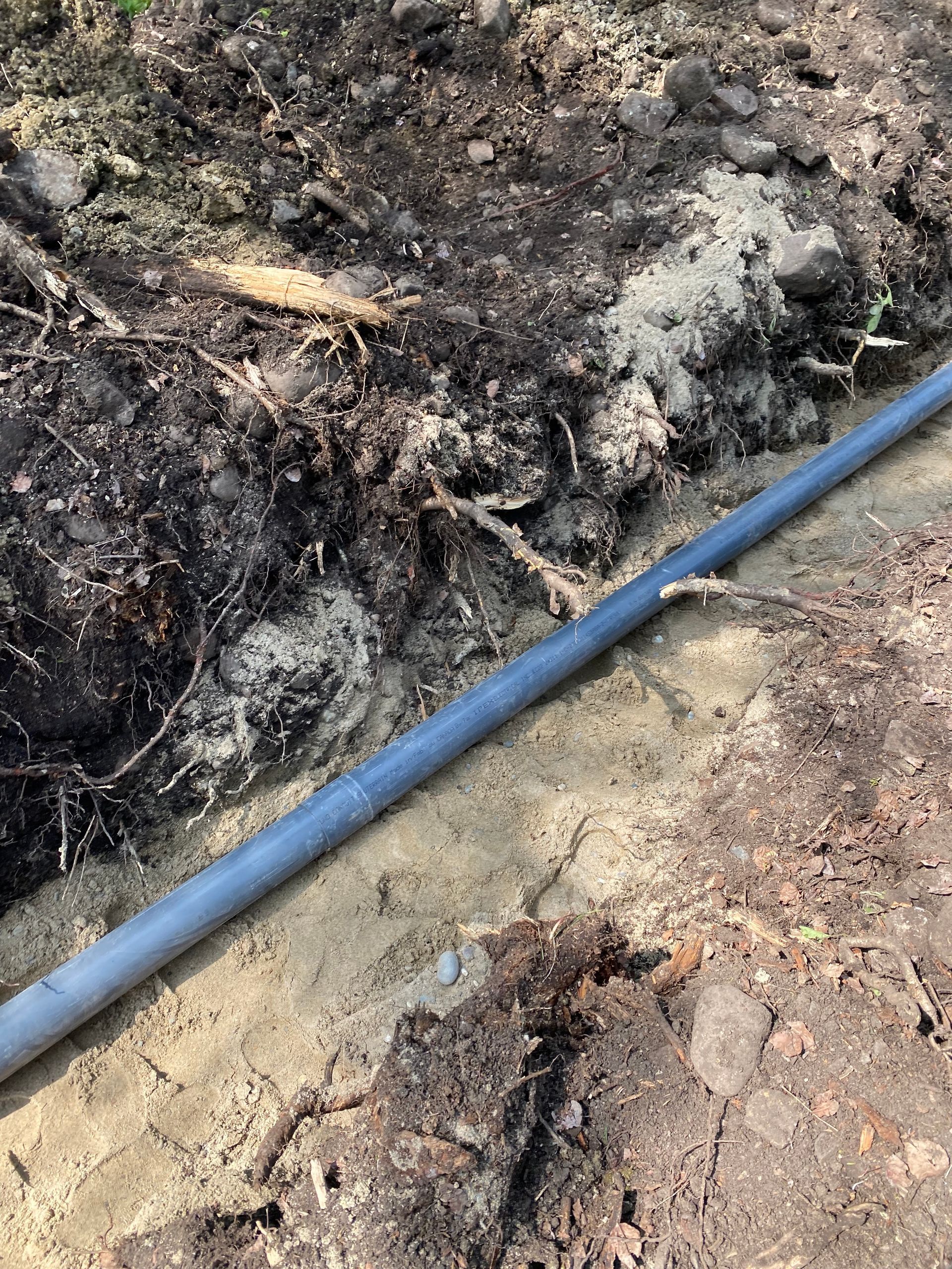 A gray pipe laid in a shallow trench filled with tan sand and surrounded by dark soil.