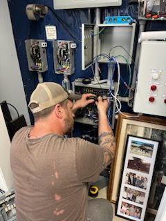 Man in cap working on electrical wiring, blue panel backdrop, picture frame visible.