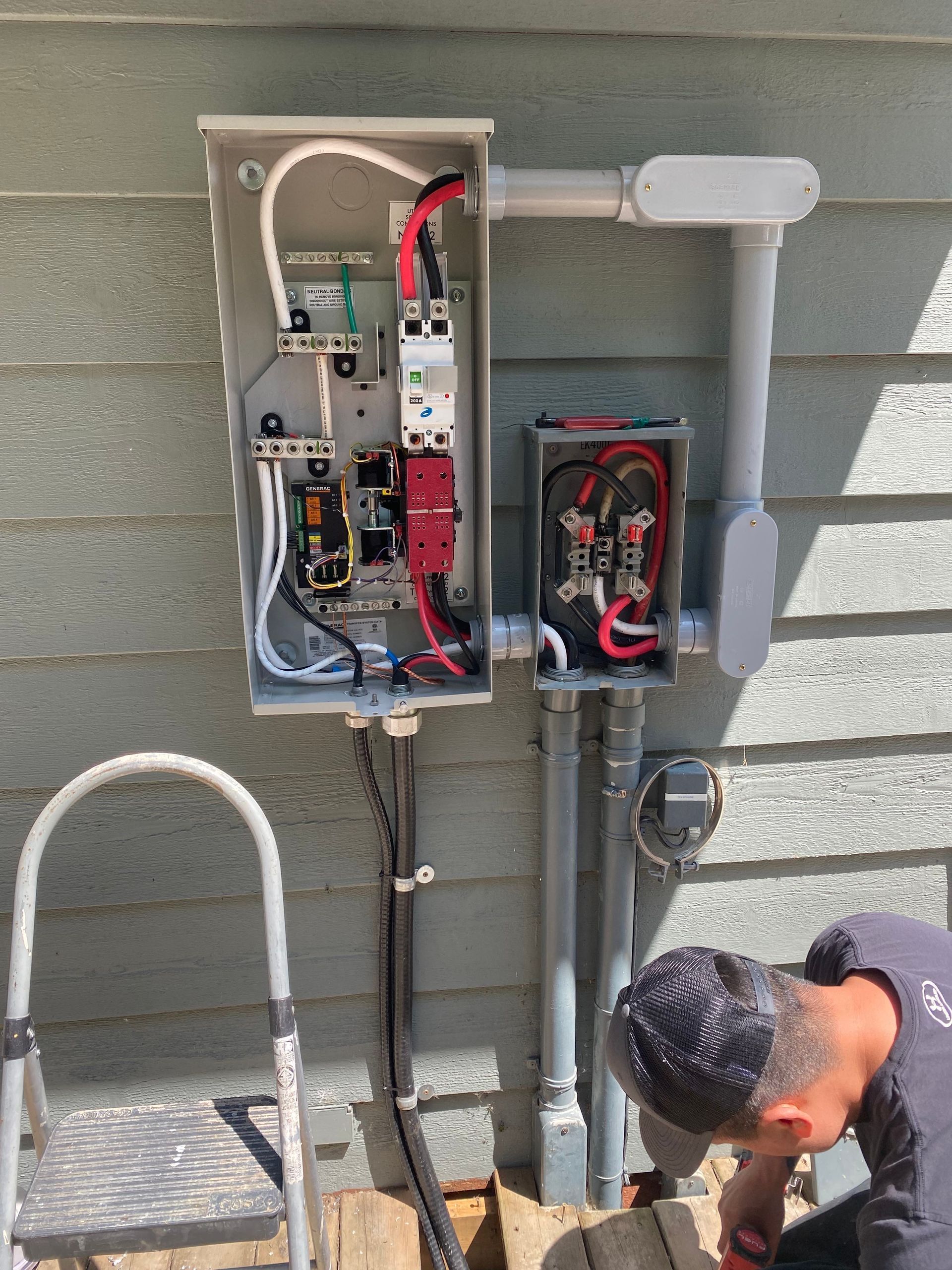 Electrician working on electrical panel attached to a building's siding. Wires, conduits, and equipment visible.