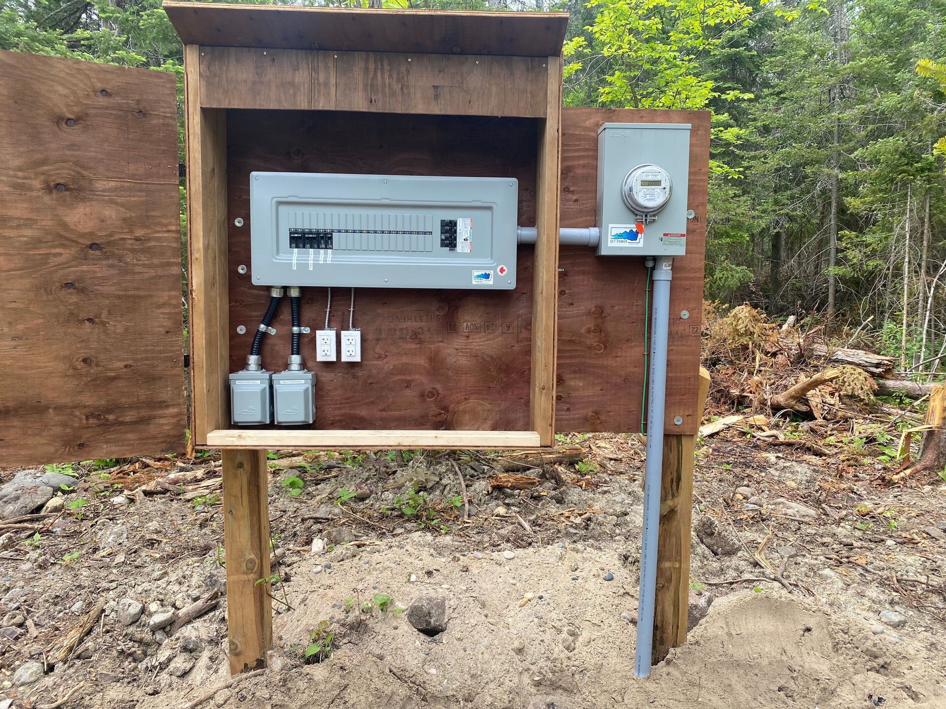 Electrical panel and meter mounted on wooden supports, outdoors. Brown plywood, gray panel, gray conduits.