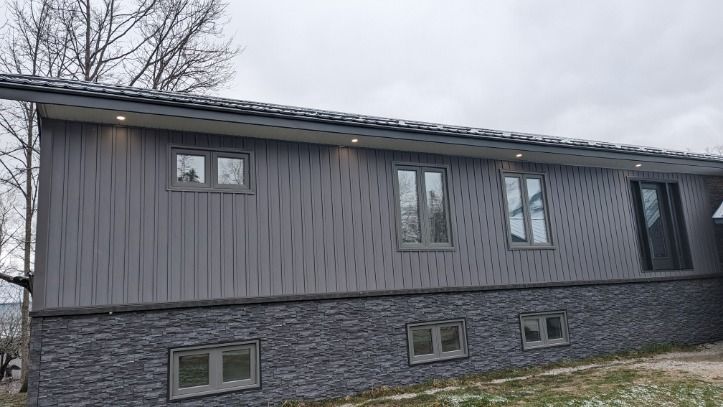 Gray house with stone base and vertical siding. Multiple windows. Cloudy day.
