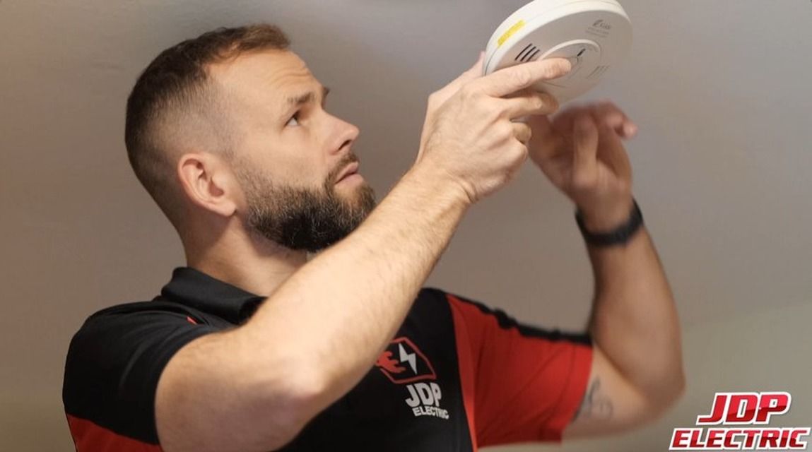 Man in black-and-red shirt installing a smoke detector on a ceiling.