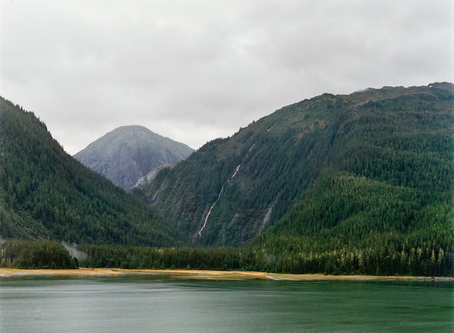 Cloudy mountain lake with forested hills and a distant peak reflected in calm water