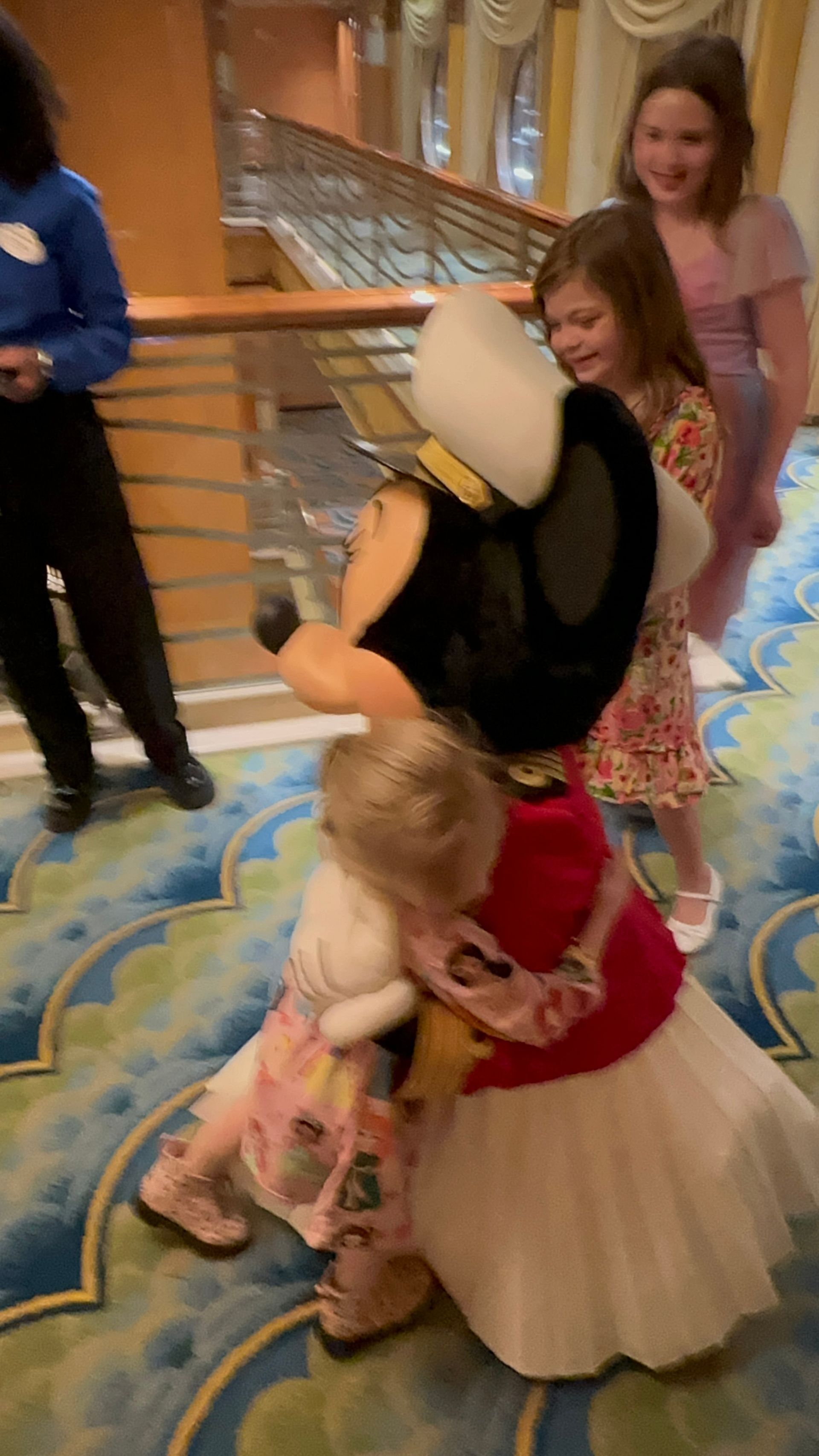 Mickey Mouse hugs a child; two other girls watch. Interior setting with green carpet.