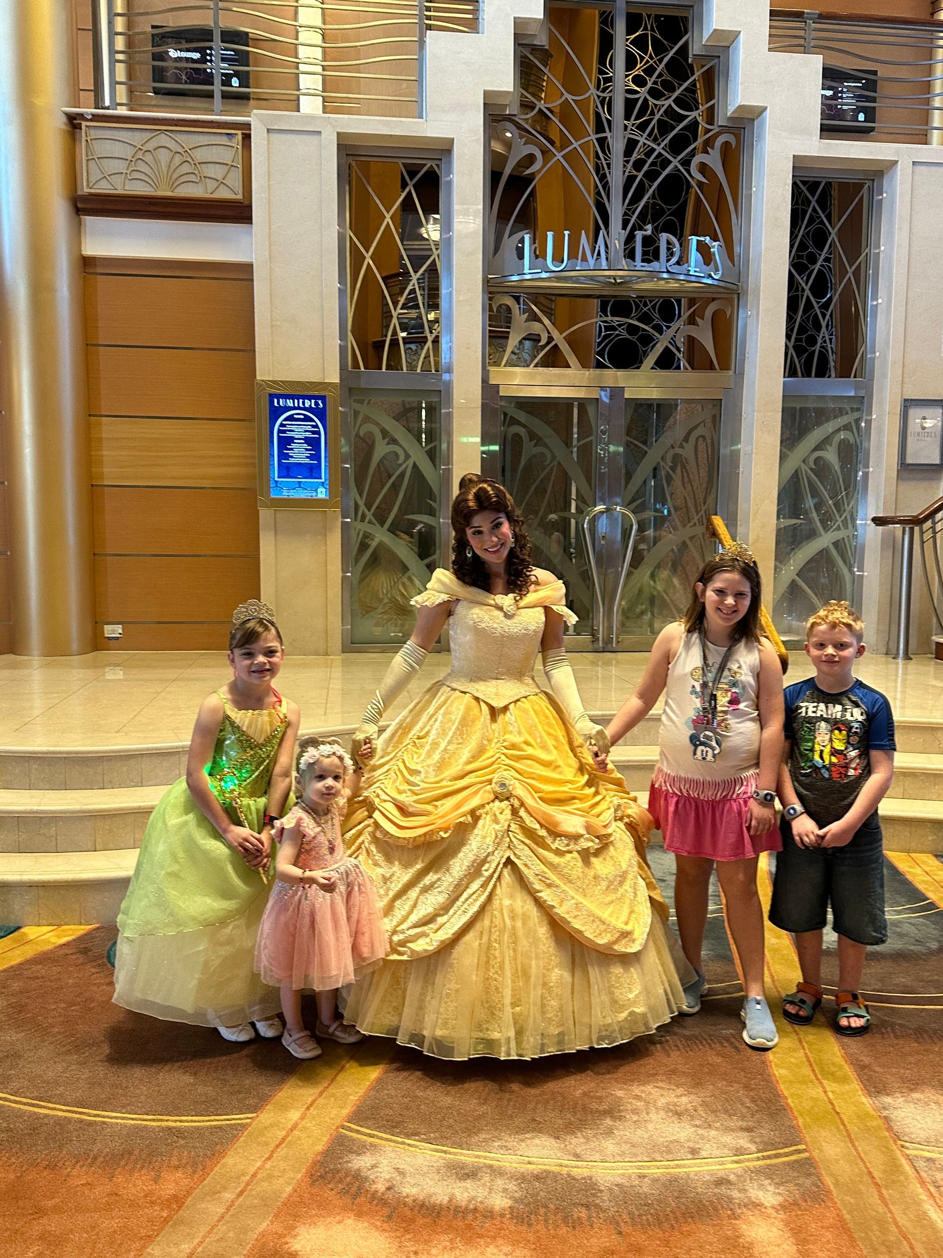 Snow-capped mountains rise above a dark forest and a calm blue body of water, under a bright blue sky.