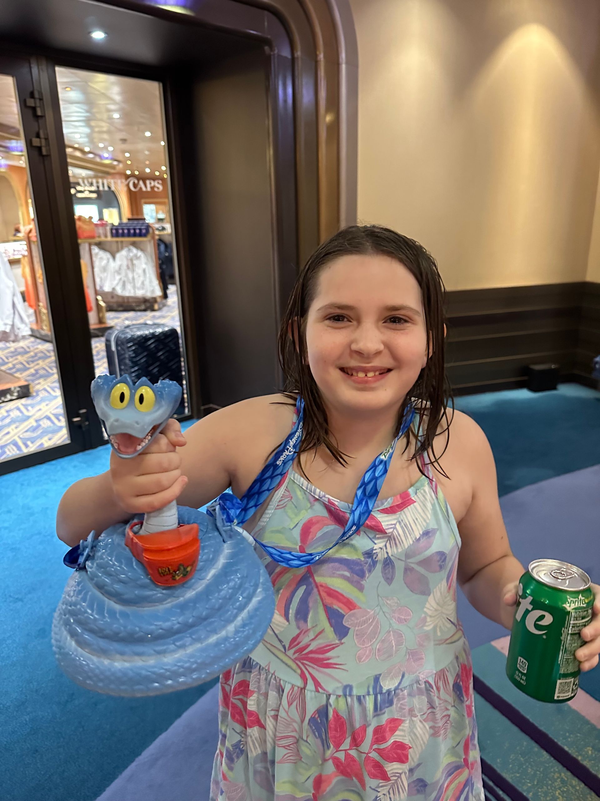 Young person with a blue snake toy and a soda can smiling. Interior with blue carpet and doorway.