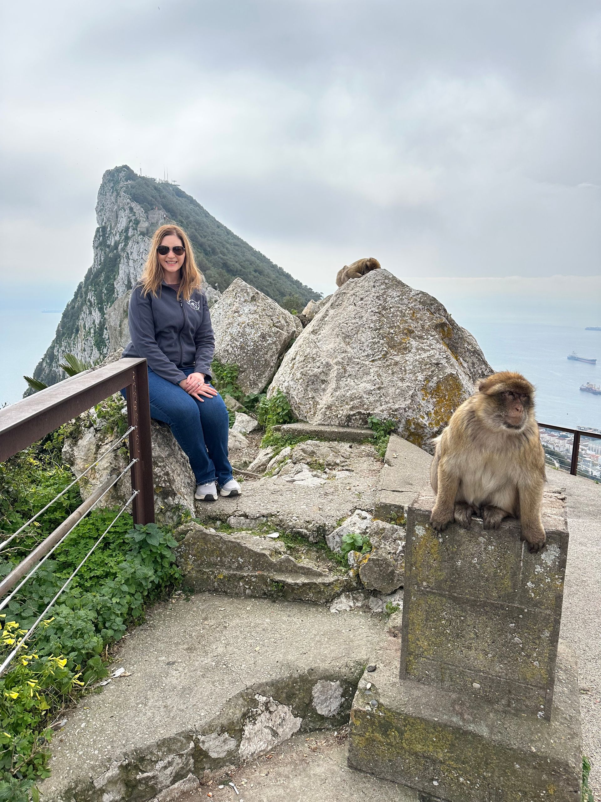 Woman sits near monkey on a rocky ledge, overlooking the sea and a large rock formation.