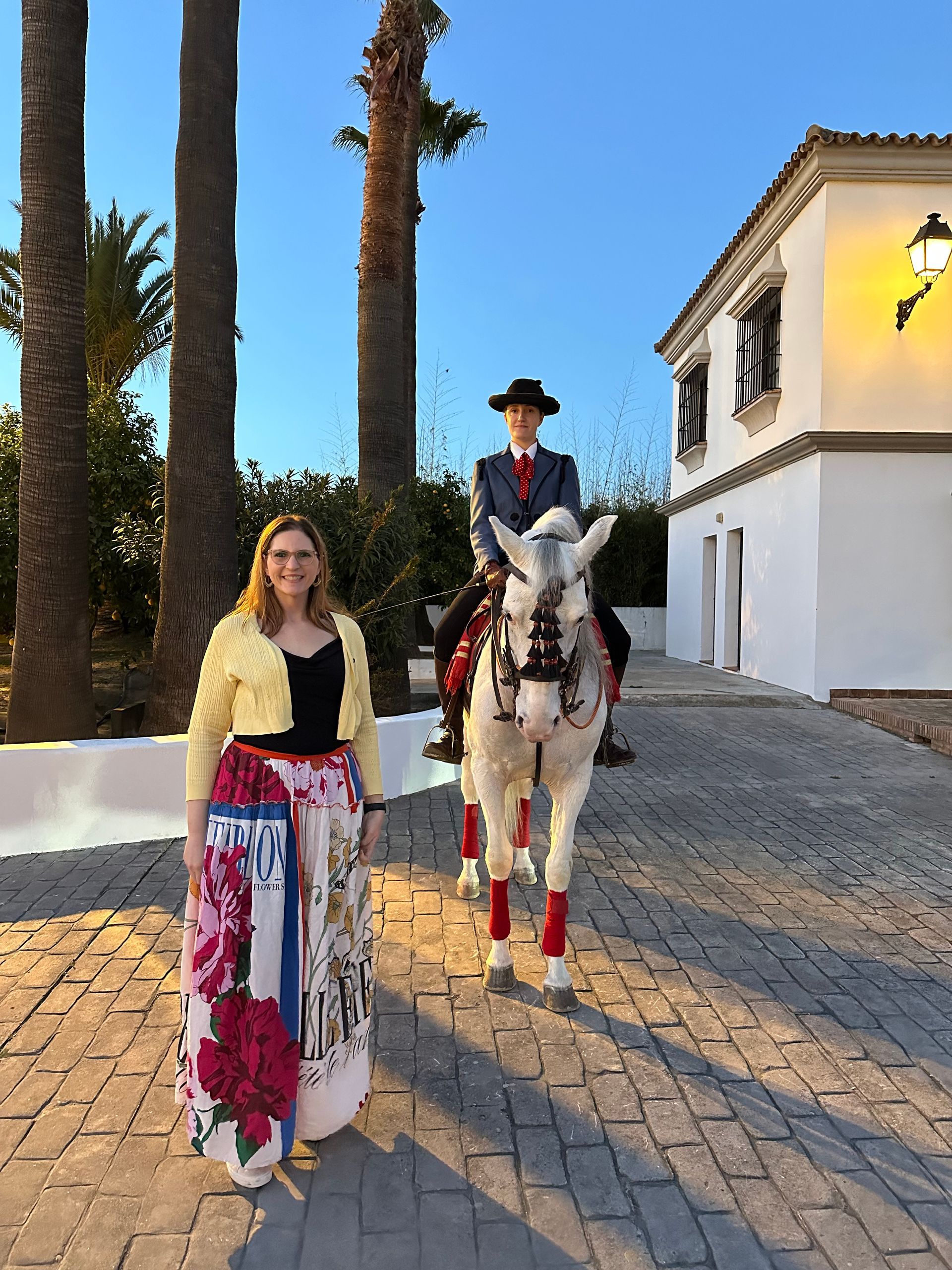 Woman beside a rider on a white horse, both in formal wear, in front of a white building with palm trees.