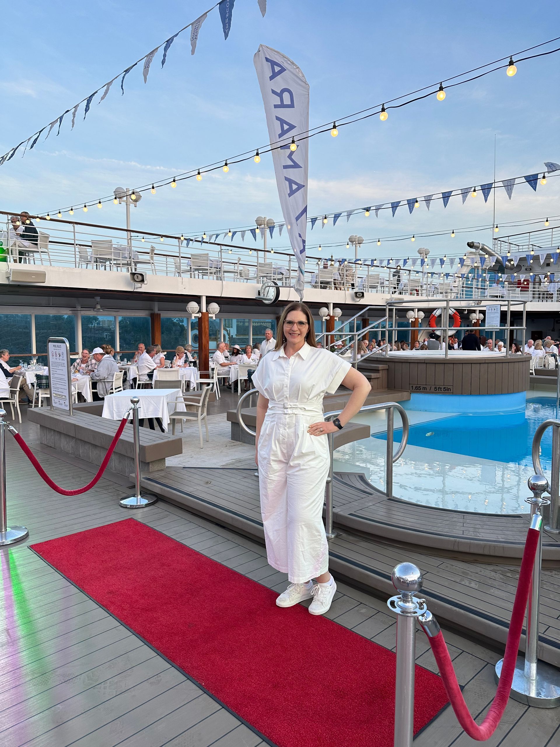 Woman in white outfit stands on red carpet on cruise ship deck.