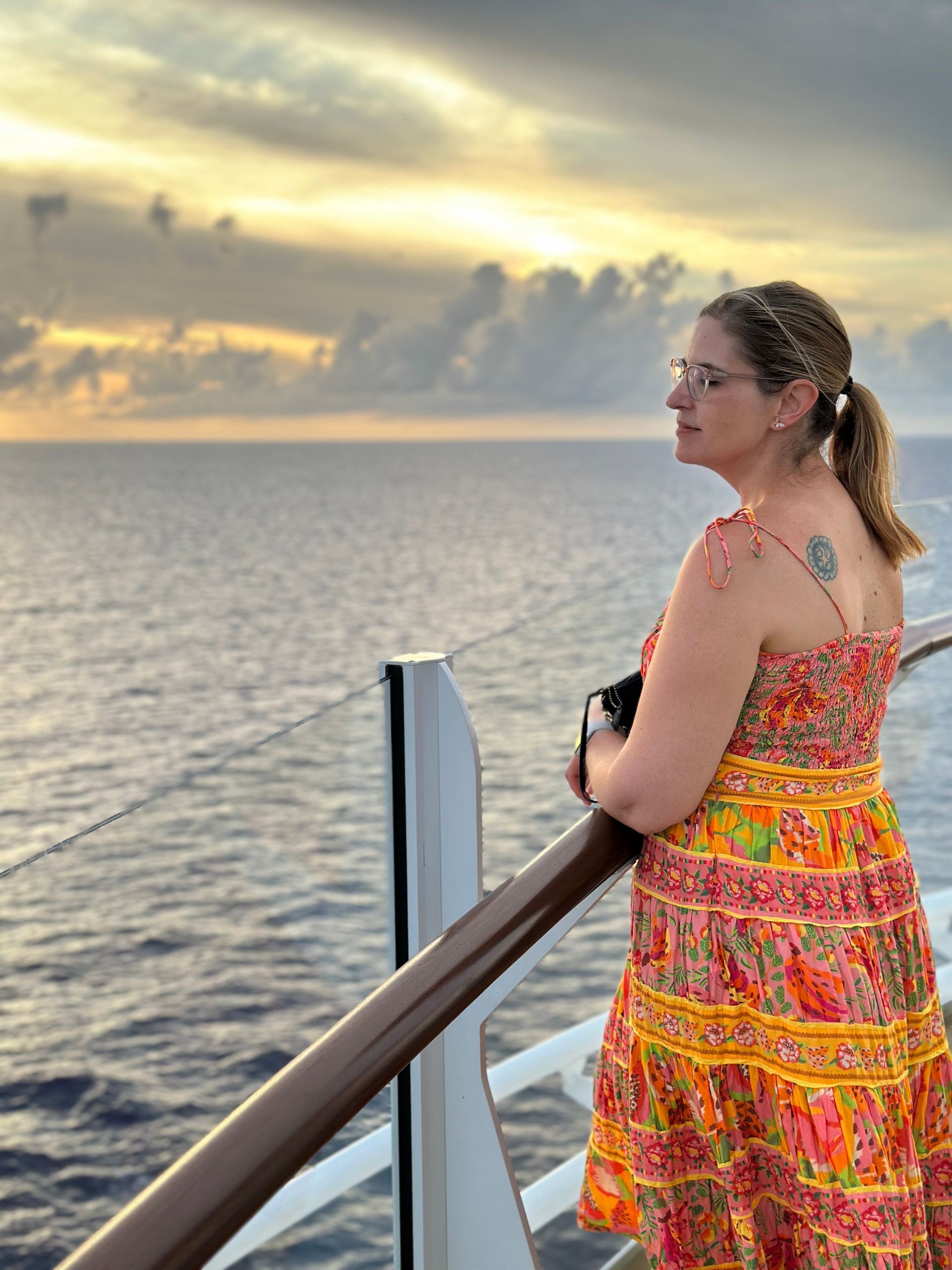 Woman in colorful dress gazes out at ocean sunset from a ship's railing.