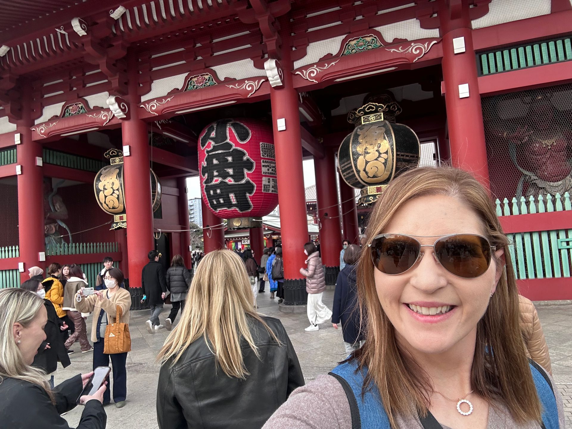 Woman in sunglasses smiles in front of a red temple with large lanterns in Japan.