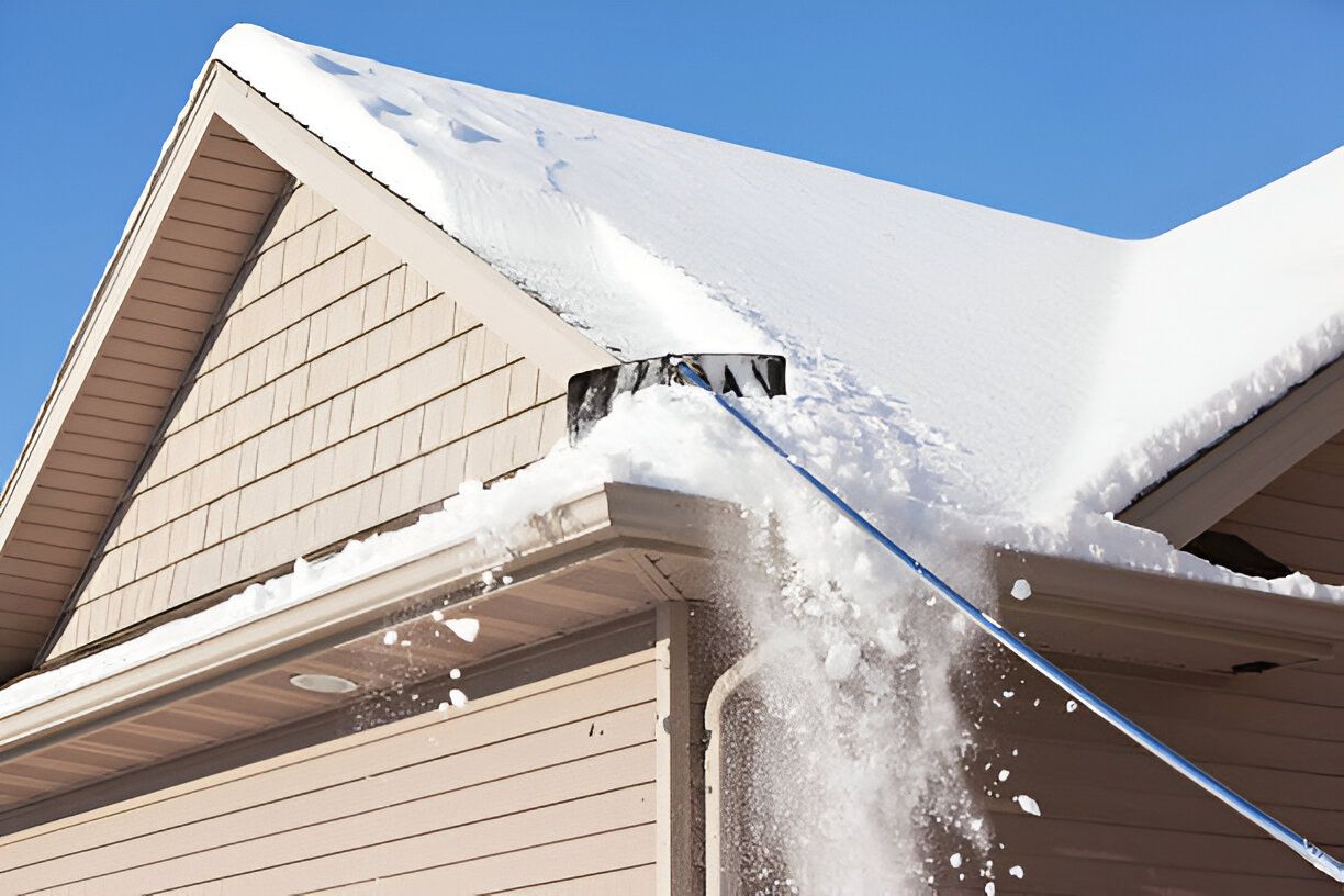 Snow being removed from a roof with a long-handled snow rake on a sunny day.