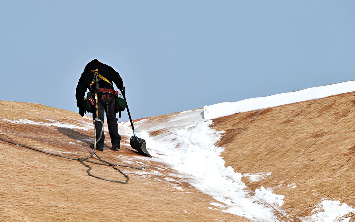 A person on a roof shoveling snow, secured by a harness and rope. Blue sky.
