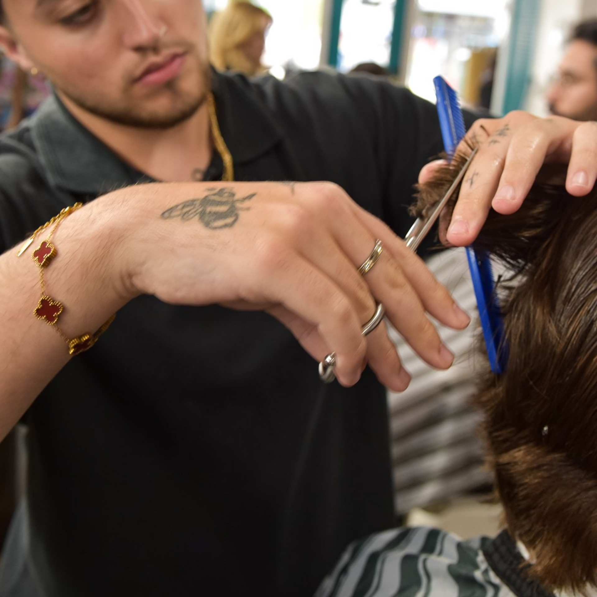Barber cutting hair with scissors and comb. Man has tattoos and jewelry, indoors.
