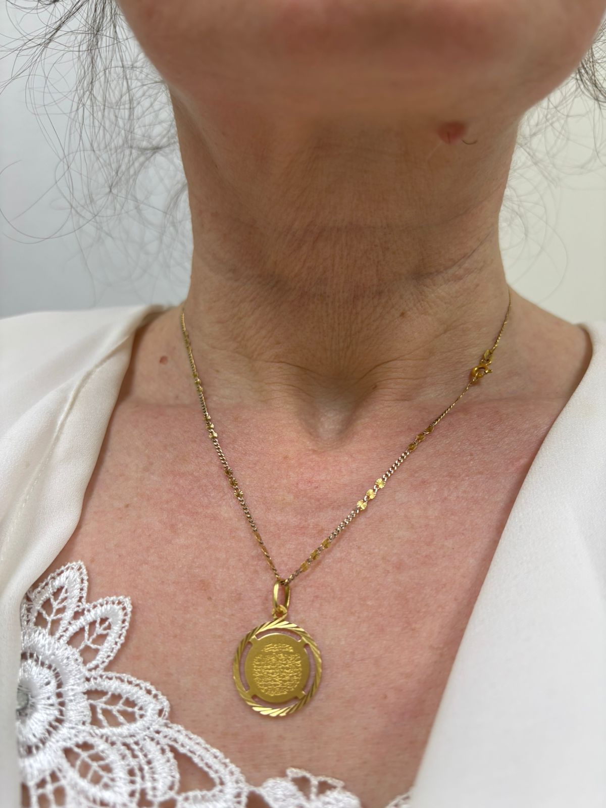 Woman wearing a gold necklace with a round pendant, against a white blouse with lace detail.