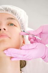 A person receiving a facial injection; hands in pink gloves, needle near cheek.