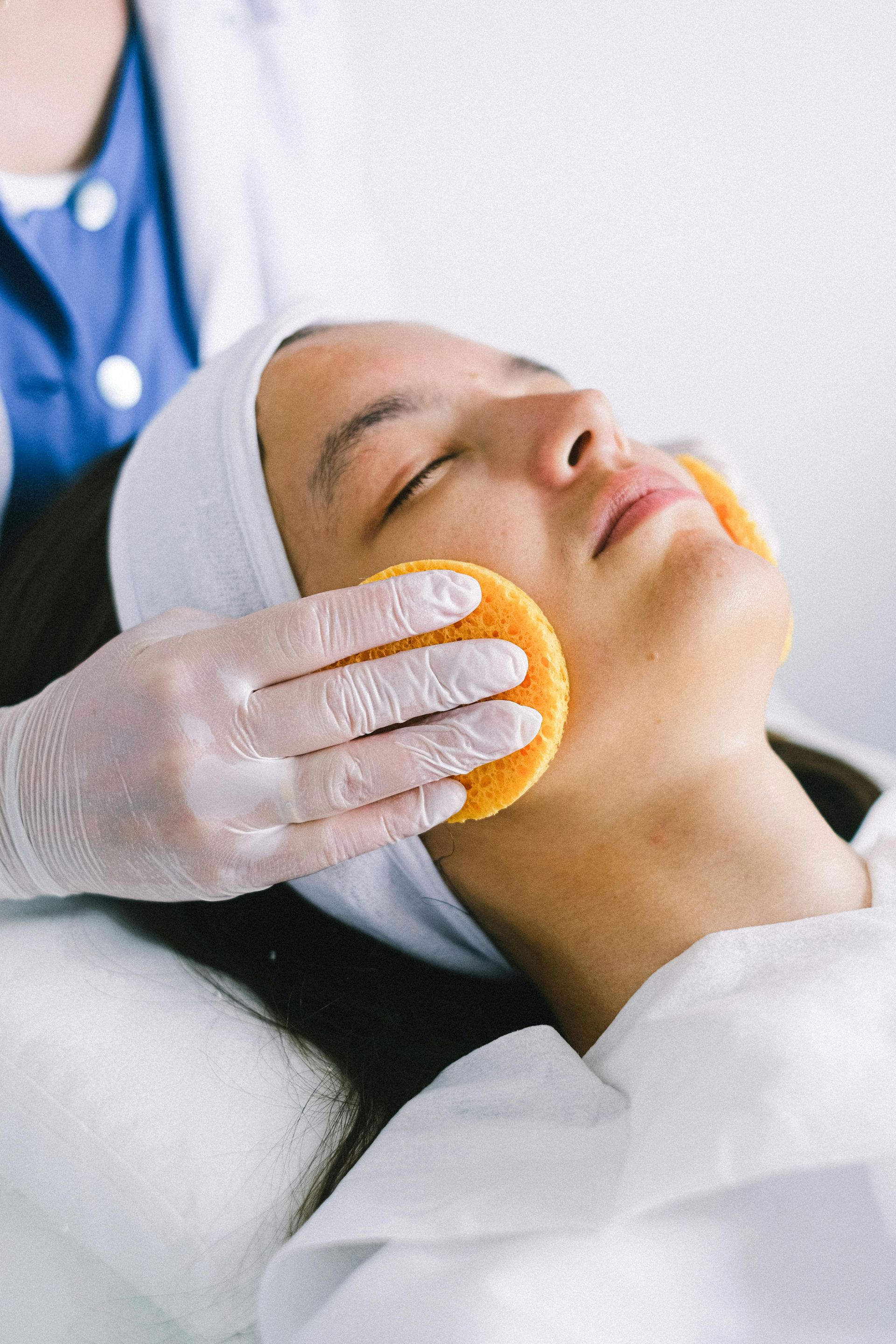 Person receiving a facial treatment; esthetician cleaning face with an orange sponge.