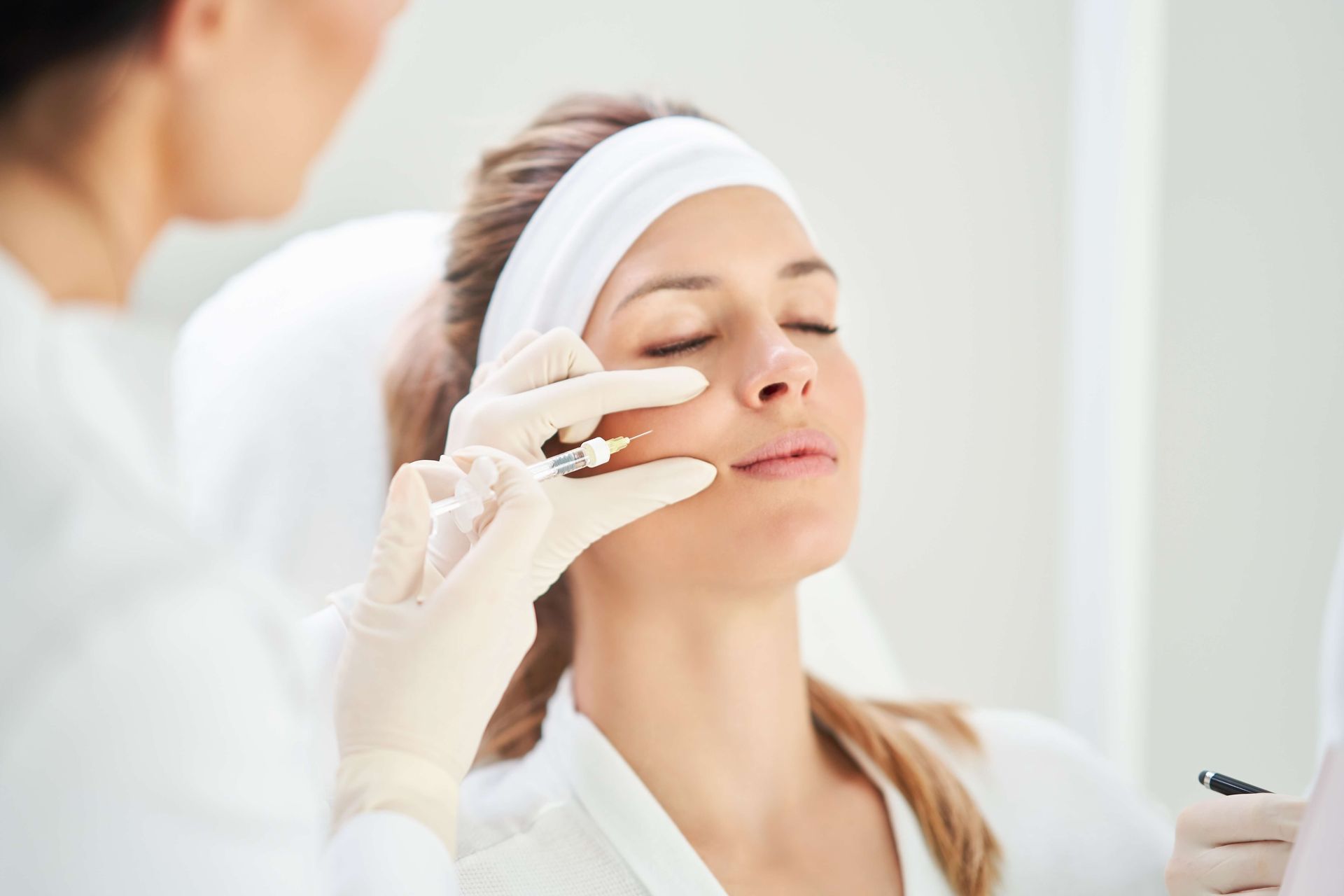 Person receiving a facial injection around the mouth. Pink gloves, syringe, and a medical setting are visible.
