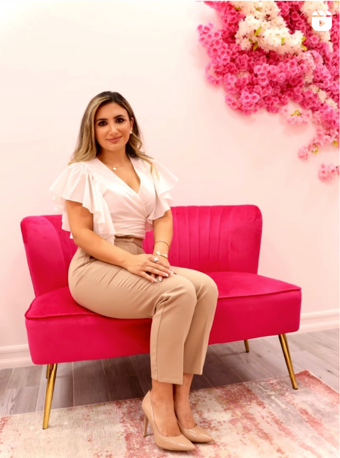 Woman sitting on a pink couch, wearing a white top and tan pants, with pink floral wall decor.