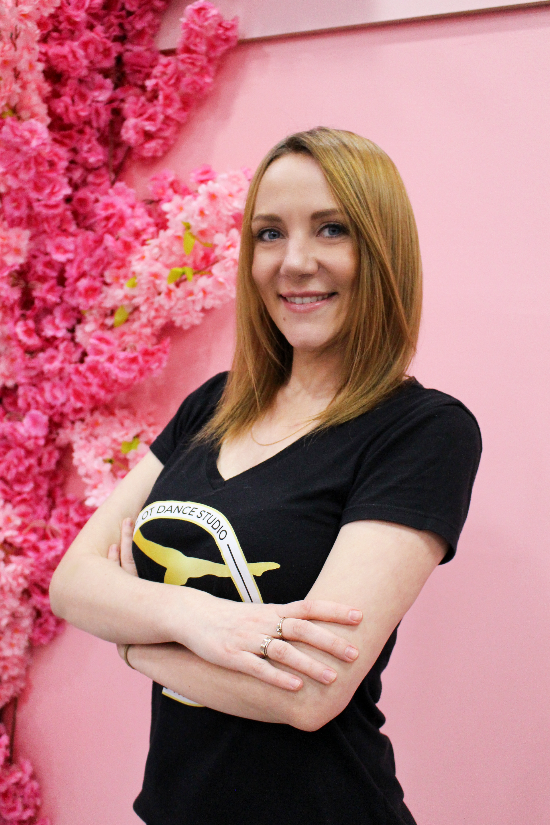 Woman with crossed arms smiling in front of pink flowers. She wears a black shirt and has light brown hair.