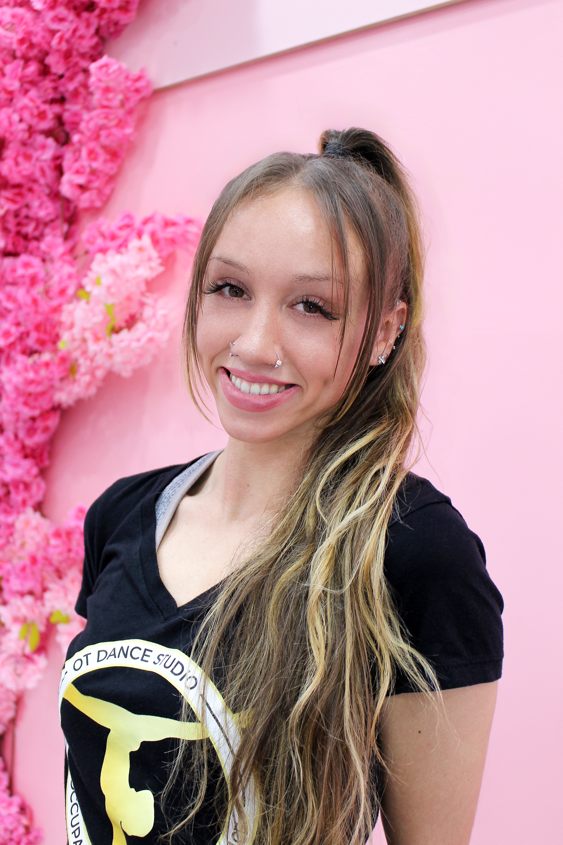 Woman with long highlighted hair smiles in front of pink flower wall. She's wearing a black shirt.